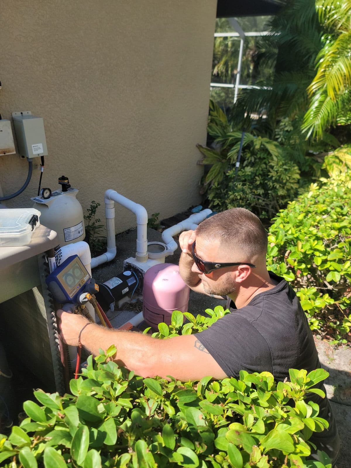 Technician diagnosing an outdoor air conditioner between shrubs