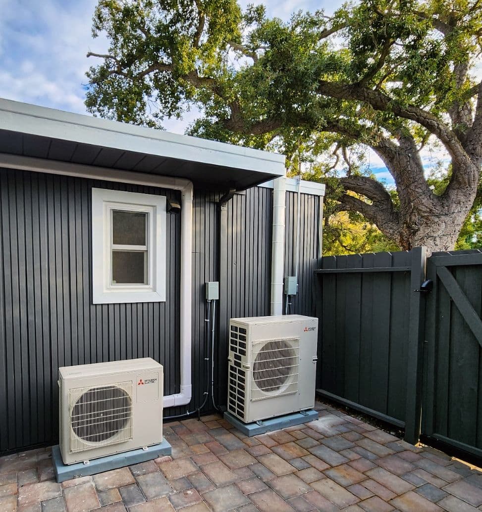 Two mini split outdoor units beside a dark painted house