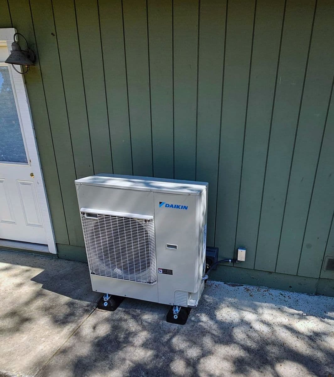 Daikin mini split outdoor unit on a pad against green siding